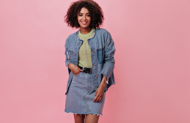 Dark-skinned woman in denim outfit looking at camera on pink background. Portrait of curly girl in