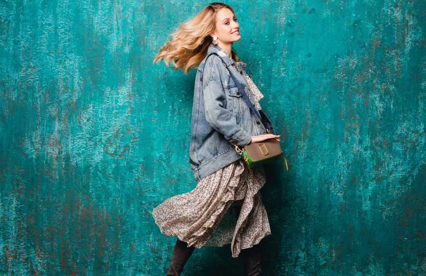sexy attractive stylish blonde woman in jeans oversize jacket walking against vintage green wall in street, autumn spring summer fashion trend, wearing silk dress, purse, urban style, smiling, happy