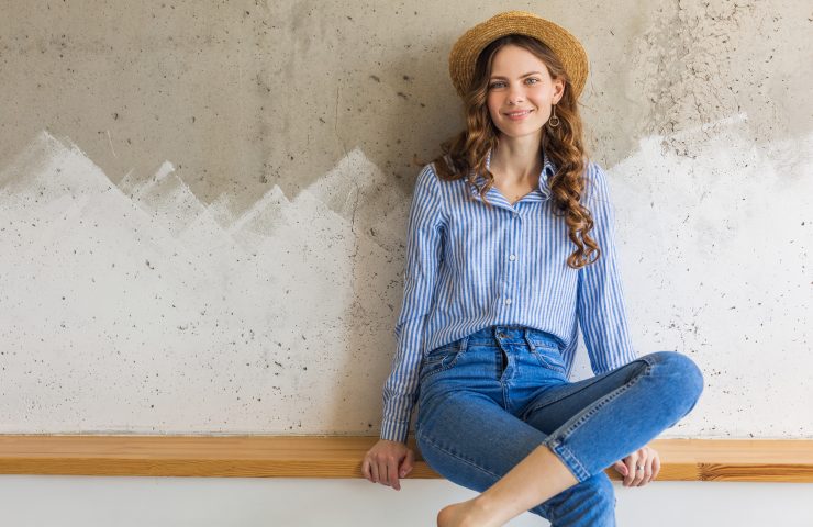 young attractive stylish woman sitting at wall background, straw hat, wearing blue cotton shirt, jeans, casual outfit, hipster style, spring summer fashion trend, smiling face, happy mood, relaxed